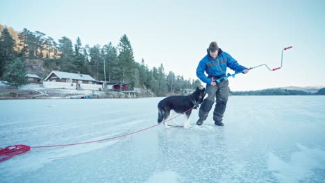 Man-With-Pet-Alaskan-Malamute-Drills-Hole-Through-Thick-Ice-With-A-Hand-Ice-Auger-For-Ice-Fishing-On-Frozen-Lake-In-Winter