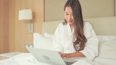 Brunette-woman,-wearing-white-bathrobe-and-sitting-on-her-bed,-using-a-laptop-and-typing-on-the-keyboard