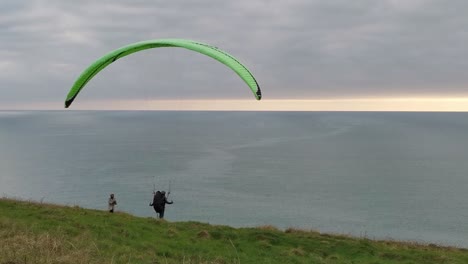 A-man-trying-to-catch-a-wind-with-his-paraglide-while-woman-is-watching-at-the-edge-of-the-cliff-overlooking-the-Atlantic-Ocean-on-a-cloudy-day-with-sunset-sun-over-horizon