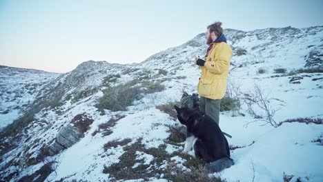 Male-Hiker-And-Alaskan-Malamute-Eating-Banana-While-Taking-A-Break-On-A-Snowy-Mountain-In-Norway