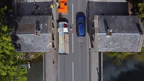 Top-down-of-an-old-stone-bridge-with-toll-buildings-incorporated,-traffic-filtering-through-with-roadworks-taking-place