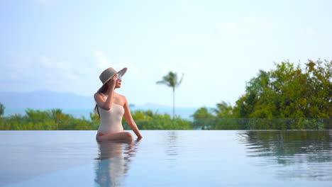 A-pretty-woman-in-a-one-piece-bathing-suit,-sunglasses,-and-sun-hat-relaxes-on-the-edge-of-an-infinity-pool