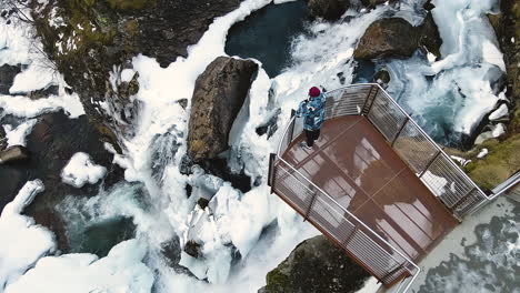 Vista-Superior-De-Un-Turista-En-La-Plataforma-De-Observación-De-Una-Cascada-Cubierta-De-Nieve-En-La-Ciudad-De-Geiranger,-Noruega-Occidental