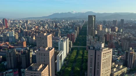 Slow-aerial-forward-flight-over-green-famous-renai-road-with-trees-surrounded-by-high-rise-buildings-and-silhouette-of-mountains-in-background