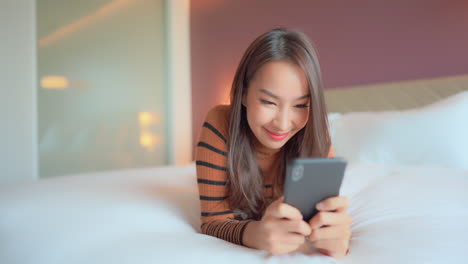 Asian-woman-holding-phone-in-both-hands-and-typing-in-messenger-while-lying-on-bed-smiling