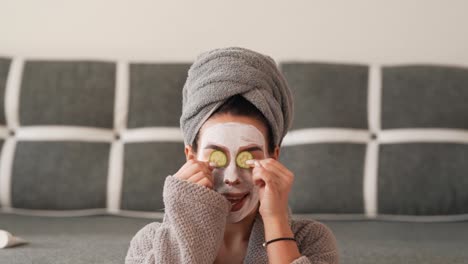 Pretty-black-haired-woman-with-a-towel-on-her-head-and-a-white-cosmetic-mask-on-the-forms-indulges-in-cucumber-slices