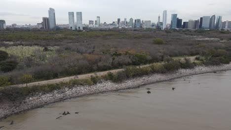 Aerial-view-of-people-cycling-on-the-unpaved-trail-along-the-river,-on-outskirts-of-Buenos-Aires