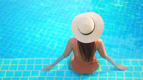 Back-view-of-unrecognizable-woman-in-orange-monokini-and-sunhat-sitting-on-the-border-of-hotel-swimming-pool-inside-water,-slowmotion-elevated-point-of-view-static