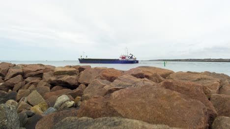 Large-blue-cargo-ship-leaving-Port-of-Liepaja-,-overcast-spring-day,-wide-revealing-shot-from-tilt-up-from-stone-pier-rocks