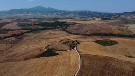 Aerial-movie-location-of-Gladiator-starring-Russell-Crowe-in-Val-d'Orcia-near-Siena,-Florence-and-Pienza-with-an-avenue-of-cypress-trees-with-harvested-wheat-crop-fields-on-panoramic-summer-hills