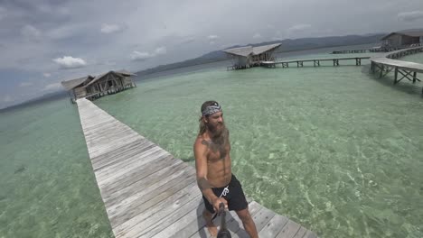 A-young,-fit-and-strong-man-with-long-hair-and-beard-is-walking-on-the-jetty-and-spins-around-to-show-the-beautiful-surrounding-of-the-luxury-resort-with-individual-bungalows-out-in-the-water