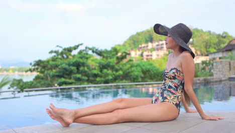 Asian-Woman-in-Tropical-Pattern-Swimwear-and-Black-Sunhat-Resting-on-the-Floor-Next-to-Swimming-Pool-Looking-Aside-at-Outdoor-Hotel-Lounge,-slow-motion-static
