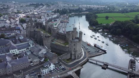 Ancient-Caernarfon-castle-Welsh-harbour-town-aerial-view-medieval-waterfront-landmark-high-lowering-shot