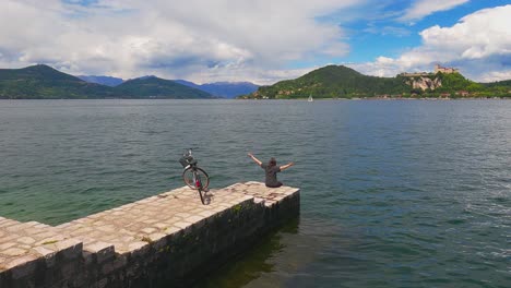 Back-view-of-carefree-happy-woman-with-arms-open-and-bicycle-on-jetty-edge-of-Maggiore-lake,-Italy