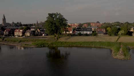 Aerial-backwards-movement-of-riverbank-with-tree-reflecting-in-the-river-IJssel-revealing-the-towers-of-Zutphen-in-the-background-on-an-overcast-day-during-sunset