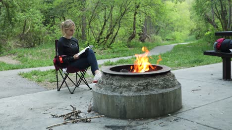 Woman-reading-book-beside-campfire-on-camping-outdoors-vacation