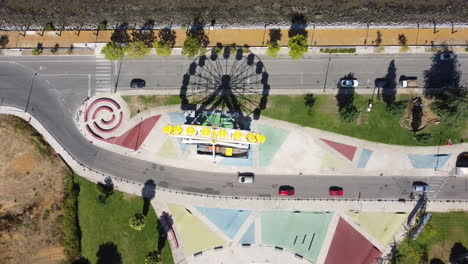 Aerial-View-Of-Ferris-Wheel-With-Shadow-At-The-Amusement-Park-In-Seixal,-Portugal-With-Cars-Passing-By-On-The-Street-At-Daytime