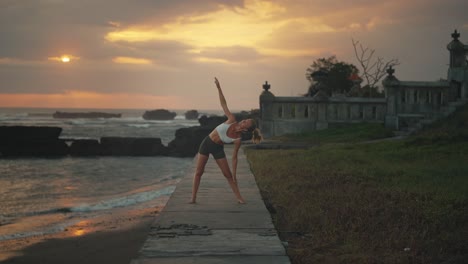 Woman-doing-Triangle-pose-with-dramatic-sunset-in-background,-Bali-coast