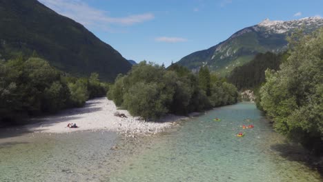 A-Group-of-Kayaks-Going-Downstream-the-Clear-Soča-River-in-Slovenia-with-a-Group-Having-a-Picnic-in-a-Dried-Up-Riverbed