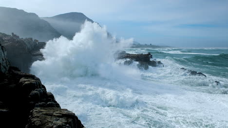Huge-wave-making-a-big-splash-as-it-crashes-into-rocks-on-rocky-coastline,-Hermanus