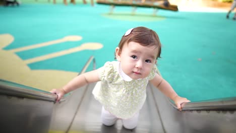 Cute-little-one-year-old-baby-Girl-standing-on-metal-slider-in-a-kid's-playground-and-staring-at-camera-lens-with-curiosity-and-astonishment,-baby-funny-facial-expression