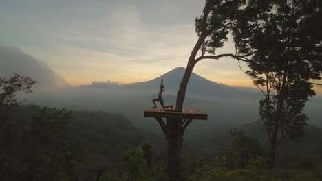 Woman-doing-warrior-pose-on-wooden-platform-with-misty-view-of-Mount-Agung,-dusk