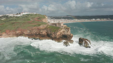 Fliegen-über-Ein-Wildes-Meer,-Das-Auf-Felsen-In-Richtung-Fort-Von-São-Miguel-Arcanjo-Mit-Einem-Regenbogen-Am-Himmel-Stürzt