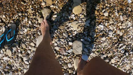 First-person-fpv-view-of-man-feet-and-legs-leaving-blue-flip-flops-and-walking-on-stony-seashore-toward-clear-sea-water