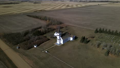 Drone-footage-of-beautiful-old-heritage-church-sitting-secluded-in-north-American-prairie-landscape-while-single-car-drives-along-straight-road-in-the-background