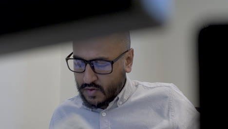 Bald-Ethnic-Minority-Entrepreneur-Turning-Head-Side-To-Read-Reading-On-Desk,-Viewed-Behind-Computer-Monitors