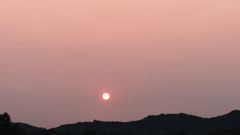 Cloudless-hazy-sunset-time-lapse-as-sun-goes-behind-distant-forested-hills,-4K