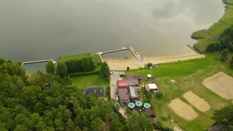 Aerial-fly-over-hotel-resort-facility-with-private-wooden-jetty-for-water-sports-like-kayaking,-canoeing-pedal-boarding,-and-fishing-boats-in-the-waterfront-of-Hartowiec-lake,-Poland