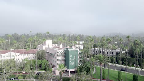 Drone-Wide-View-of-Beverly-Hills-Hotel,-Palms-and-Front-Entrance-Sign-on-a-Foggy-Day,-establishing-shot