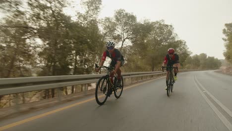 Dos-Ciclistas-De-Carretera-Profesionales-Cabalgando-Por-La-Carretera-Vacía-Del-Bosque-Y-El-Sol-Brilla-En-La-Cámara
