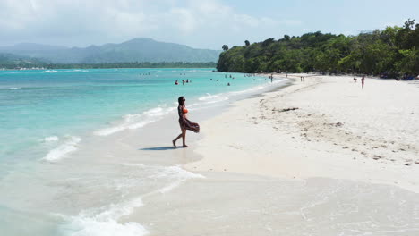 Young-woman-strolls-on-beach-showcasing-pristine-Caribbean-waters,-aerial-dolly-forward