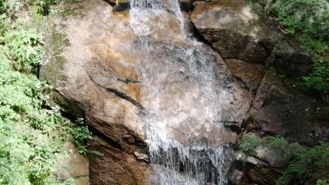 Clear-water-streams-down-rocks-and-creates-a-small-waterfall-amongst-a-forest,-as-camera-tilts