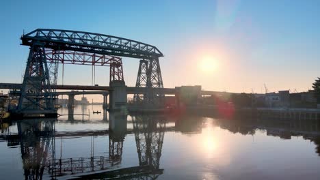 Low-level-aerial-push-out-shot-of-ferry-bridge-Nicolas-Avellaneda-over-the-Matanza-River-during-sunrise-with-sun-flare