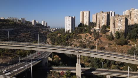 Flying-straight-through-a-complex-system-of-roads,-intersections-and-bends-in-the-hills-of-Haifa,-with-tall-buildings-surrounded-by-trees-and-a-horizon-with-clear-skies-on-a-sunny-day,-Israel