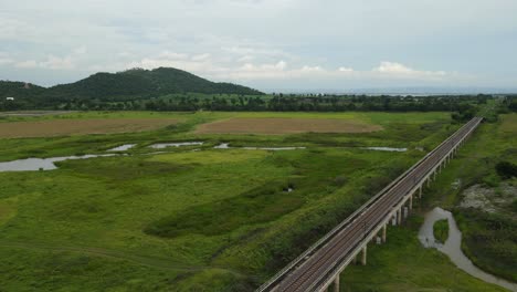 Angled-aerial-footage-towards-the-horizon-with-hills-trees,-farmland,-and-the-elevated-railway,-Muak-Klek,-Saraburi,-Thailand