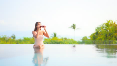 A-pretty-young-woman-wearing-a-sexy-one-shoulder-bathing-suit-sitting-on-the-edge-of-an-infinity-pool-slips-her-sunglasses-on