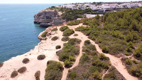 Benagil-Cave,-Algarve,-Portugal---Aerial-Drone-View-of-the-village-and-coastline,-entering-the-cave-with-tourists,-boat-and-kayaks