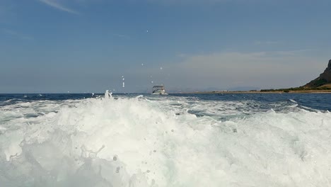 Low-angle-water-surface-pov-of-boat-wake-trail-with-Riserva-dello-Zingaro-natural-reserve-of-Sicily-in-background