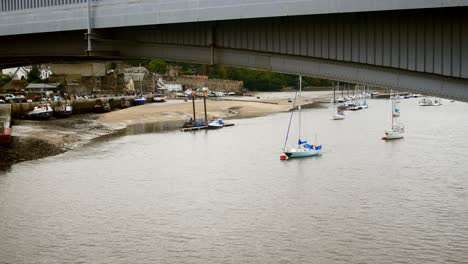 Small-sailboats-floating-under-metal-iron-arched-bridge-girder-on-scenic-fishing-town-coast