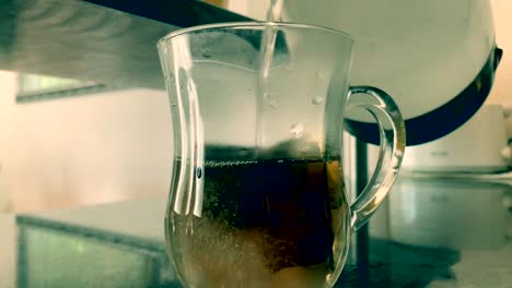 Pouring-boiling-water-from-a-glass-pitcher-into-a-cup-with-teabags-to-prepare-the-tea-beverage,-close-up-still-shot