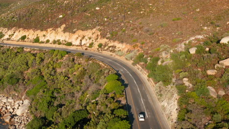 White-Car-Driving-In-Curvy-Cliffside-Highway-Of-Victoria-Road-In-Oudekraal,-South-Africa---aerial-shot