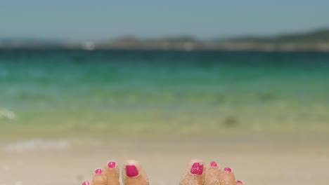 A-close-up-POV-shot-of-the-feet-of-a-young-female-sunbathing-on-a-beach,-tilting-up-to-reveal-the-beautiful-tranquil-view-of-the-surrounding-coastline-and-blue-ocean-waters