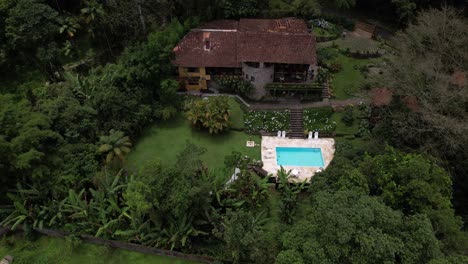 Descending-aerial-approach-of-lush-tropical-garden-greenery-with-swimming-pool-on-a-mountain-side-with-picturesque-stone-hotel-building