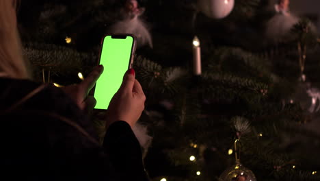 Mujer-Frente-Al-árbol-De-Navidad-Sostiene-Un-Teléfono-Inteligente-De-Pantalla-Verde,-Ots