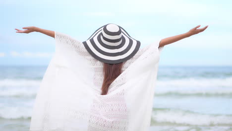Back-to-the-camera,-a-young-fit-healthy-woman-in-a-white-lacey-coverup-and-big-floppy-hat-raises-her-arms-in-joy-as-waves-from-the-ocean-roll-in