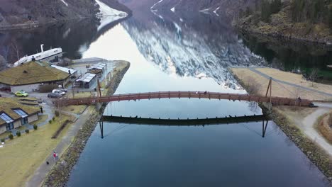 Bridge-crossing-river-in-Gudvangen-Norway---Aerial-slowly-moving-sideways-with-tourists-crossing-bridge---Beautiful-snowy-mountain-reflections-in-calm-sea-surface